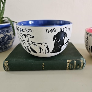 Puede incluir: Cuenco de cer&aacute;mica blanco con borde interior azul, con ilustraciones negras de perros y el texto "Dog Person". El cuenco est&aacute; sobre un libro verde.