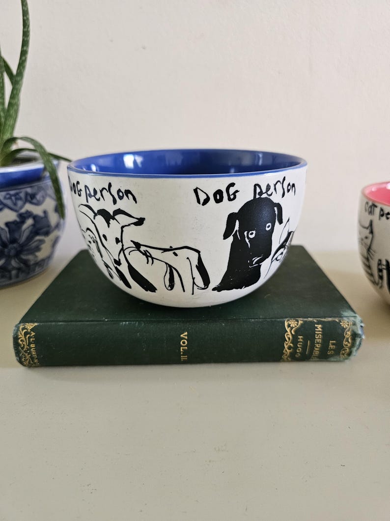 Puede incluir: Cuenco de cer&aacute;mica blanco con borde interior azul, con ilustraciones negras de perros y el texto "Dog Person". El cuenco est&aacute; sobre un libro verde.