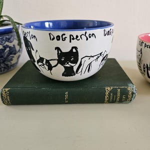 Puede incluir: Cuenco de cer&aacute;mica blanco con borde interior azul, con ilustraciones negras de perros y las palabras "Dog person". El cuenco est&aacute; sobre un libro verde.