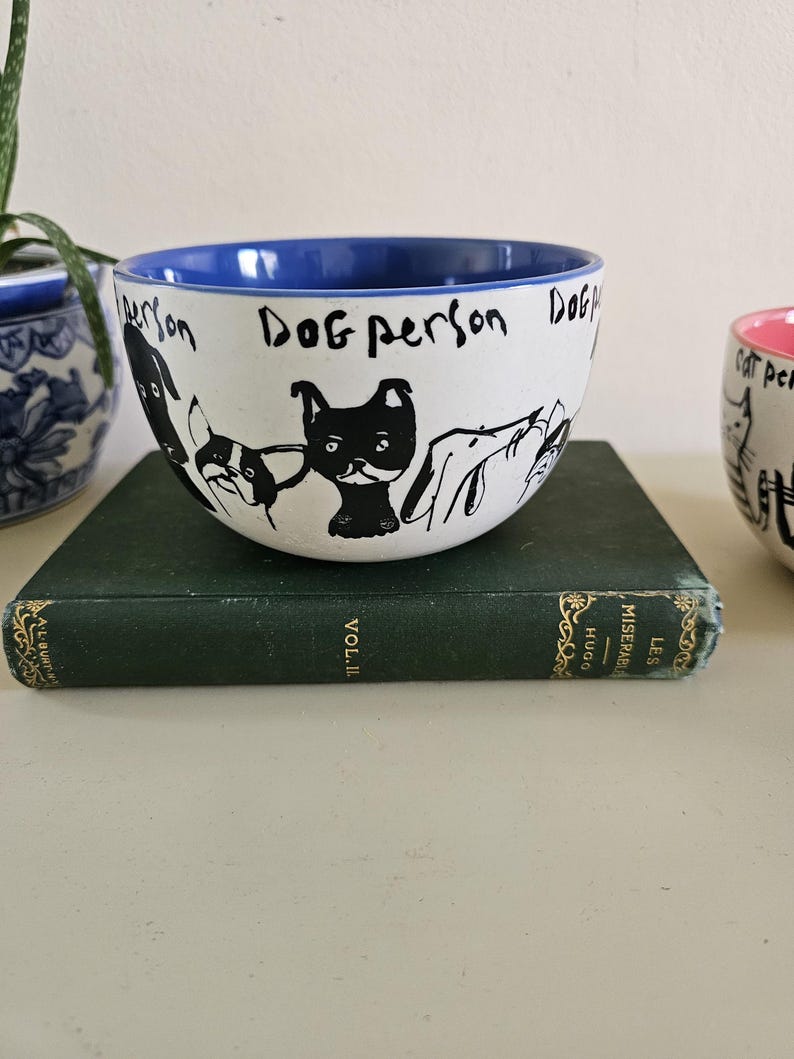 Puede incluir: Cuenco de cer&aacute;mica blanco con borde interior azul, con ilustraciones negras de perros y las palabras "Dog person". El cuenco est&aacute; sobre un libro verde.