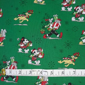 Op de afbeelding: Groene stof met een herhalend patroon van Mickey Mouse, Minnie Mouse, Donald Duck en Pluto die kerstcadeaus dragen. De personages dragen rood-wit geruite outfits en zijn omringd door sneeuwvlokken. De stof heeft de tekst "MADE IN CHINA" erop gedrukt.