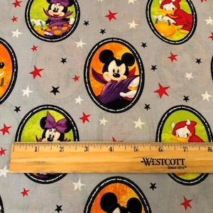 Op de afbeelding: Een grijze stof met een herhalend patroon van kleurrijke cartoonfiguren in ovale frames. De personages zijn Mickey Mouse, Minnie Mouse, Donald Duck en Goofy. De stof is versierd met rode, witte en zwarte sterren. Een houten liniaal ligt op de stof voor schaal.