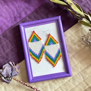 May include: A pair of rainbow-colored beaded earrings with white beads in the center, displayed in a purple frame on a patterned fabric background.