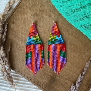Może przedstawiać: Para kolorowych kolczyków z koralików w geometryczny wzór. Kolczyki są długie i zwisające, z frędzlami z koralików na dole. Koraliki są w odcieniach czerwonego, pomarańczowego, żółtego, zielonego, niebieskiego i fioletowego.