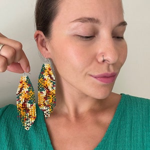 May include: A pair of long, beaded earrings with a colourful geometric pattern in shades of yellow, green, red, and white. The earrings are hanging from the ears of a person wearing a green shirt.