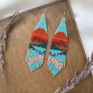 Op de afbeelding: Een paar lange, geperde oorbellen met een zuidwestelijk ontwerp. De oorbellen zijn gemaakt van turquoise, oranje, bruine en gouden kralen. De oorbellen hangen aan zilveren haakjes.