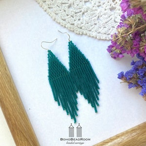 May include: A pair of long, teal blue beaded earrings with silver hooks. The earrings are hanging from a white surface with a crocheted doily and dried flowers in the background. The text "BOHOBEADROOM beaded earrings" is at the bottom of the image.