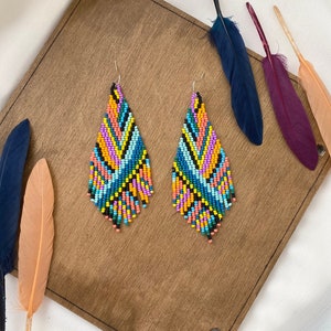 Könnte beinhalten: Ein Paar bunte, geperlte Ohrringe mit geometrischem Muster. Die Ohrringe sind aus kleinen, bunten Perlen gefertigt und haben eine Tropfenform. Die Ohrringe werden auf einer Holzoberfläche mit Federn präsentiert.