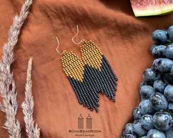May include: Gold and black beaded earrings with a fringe design. The earrings are hanging from silver hooks. The earrings are photographed on a brown fabric background.