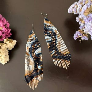 May include: A pair of beaded earrings with a butterfly wing design. The earrings feature a combination of navy blue, white, tan, and light blue beads. The earrings are long and dangle, with a silver hook.