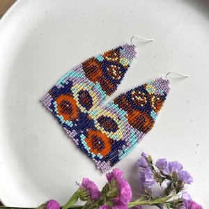 May include: A pair of long, colorful beaded earrings with a geometric pattern in shades of purple, blue, orange, and yellow. The earrings are hanging on a white plate with a floral arrangement in the foreground.