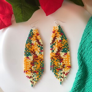 May include: A pair of long, beaded earrings with a colourful, geometric pattern in shades of yellow, orange, green, red, and white. The earrings are made of small, round beads and have a teardrop shape.