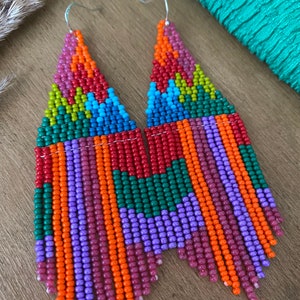 May include: A pair of long, colourful beaded earrings with a geometric pattern. The earrings are made with small, colourful beads in shades of orange, red, green, purple, and blue. The earrings are hanging from silver hooks.