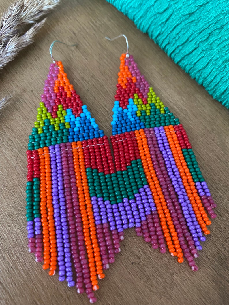 May include: A pair of long, colourful beaded earrings with a geometric pattern. The earrings are made with small, colourful beads in shades of orange, red, green, purple, and blue. The earrings are hanging from silver hooks.