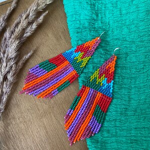 May include: A pair of colourful beaded earrings with a geometric pattern. The earrings are long and dangle, with a mix of red, orange, yellow, green, blue, and purple beads.