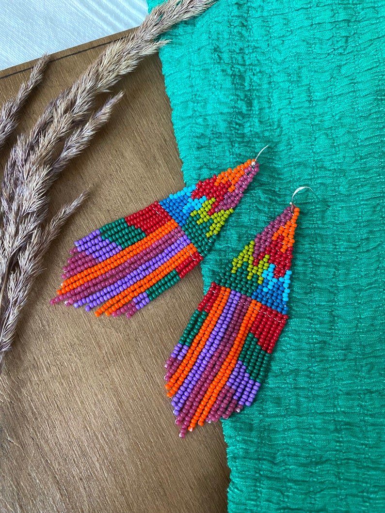 May include: A pair of colourful beaded earrings with a geometric pattern. The earrings are long and dangle, with a mix of red, orange, yellow, green, blue, and purple beads.