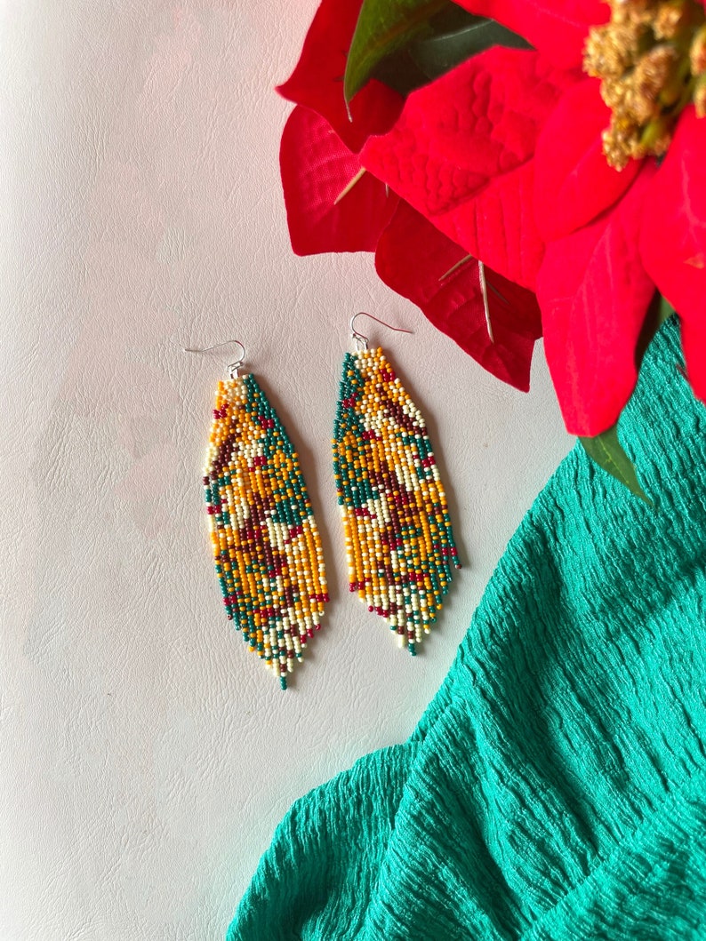 May include: A pair of long, beaded earrings with a geometric pattern in shades of green, yellow, red, and white. The earrings are hanging from silver hooks.