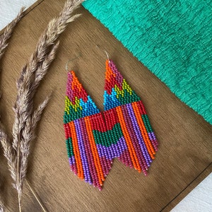 May include: A pair of long, colourful beaded earrings with a geometric pattern. The earrings are made with small, colourful beads in shades of red, orange, yellow, green, blue, purple, and pink. The earrings are hanging from silver hooks.