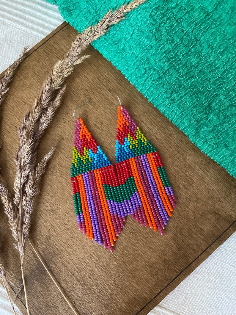May include: A pair of long, colourful beaded earrings with a geometric pattern. The earrings are made with small, colourful beads in shades of red, orange, yellow, green, blue, purple, and pink. The earrings are hanging from silver hooks.