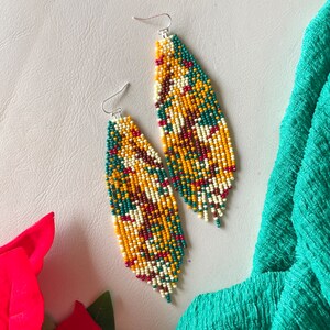 May include: A pair of long, beaded earrings with a geometric pattern in shades of yellow, green, white, and red. The earrings are made with small, seed beads and have a fringe-like design.