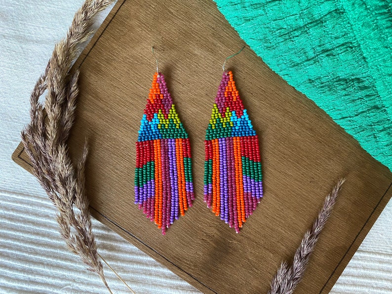 May include: A pair of colourful beaded earrings with a geometric pattern. The earrings are long and dangle, with a fringe of beads at the bottom. The beads are in shades of red, orange, yellow, green, blue, and purple.