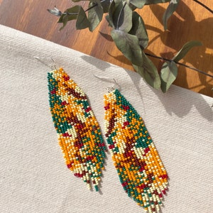 May include: A pair of long, beaded earrings with a geometric pattern in shades of green, orange, red, and white. The earrings are hanging from silver hooks.