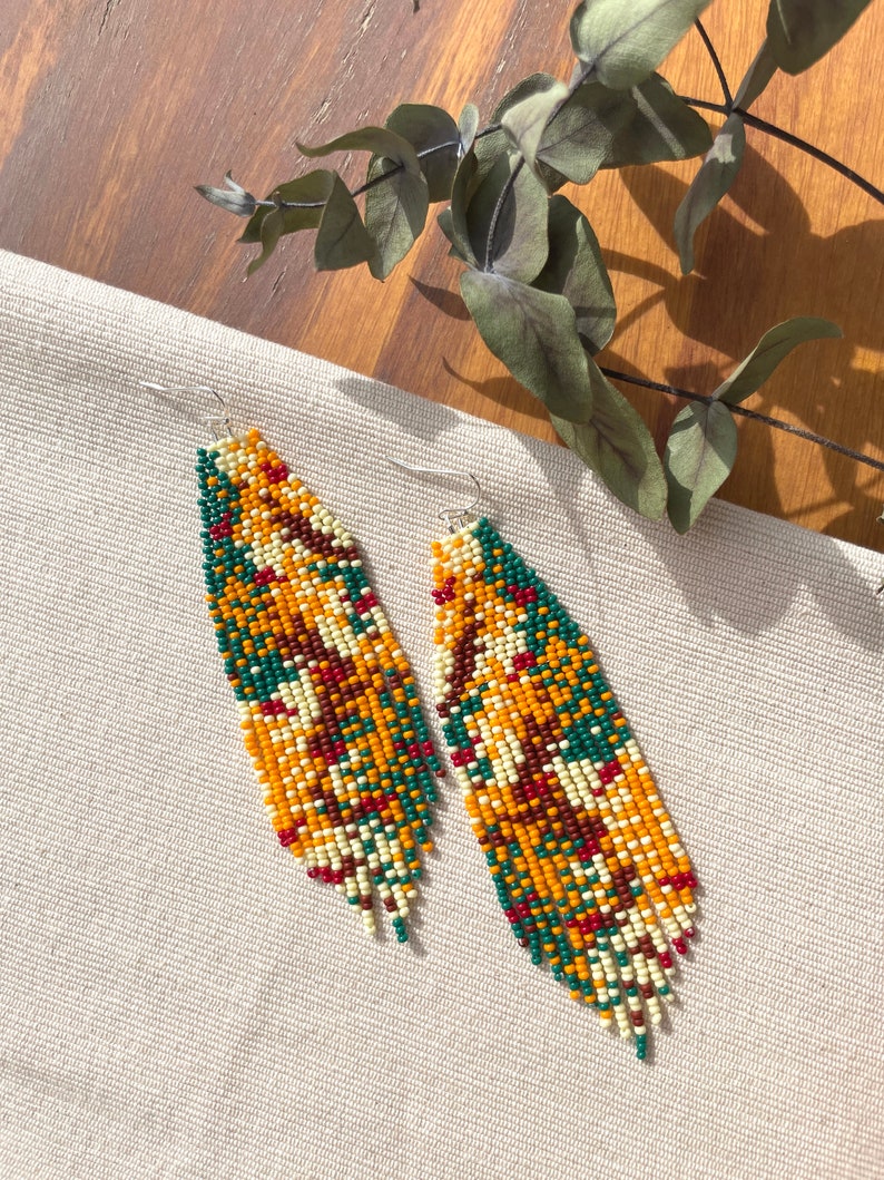 May include: A pair of long, beaded earrings with a geometric pattern in shades of green, orange, red, and white. The earrings are hanging from silver hooks.