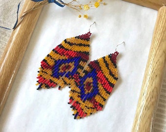 May include: A pair of beaded earrings with a geometric pattern in shades of red, orange, yellow, blue, and black. The earrings are hanging from silver hooks.