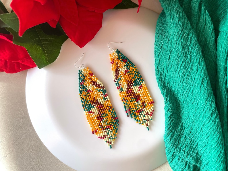 May include: A pair of long, beaded earrings with a colourful, geometric pattern in shades of yellow, green, red, and white. The earrings are hanging from silver hooks.