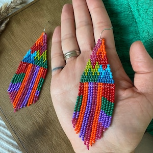 May include: A pair of colourful beaded earrings with a geometric pattern and fringe. The earrings are made with red, orange, yellow, green, blue, and purple beads.