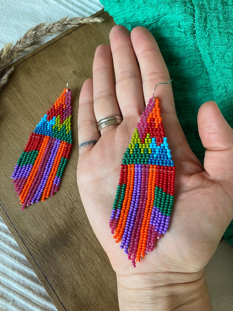 May include: A pair of colourful beaded earrings with a geometric pattern and fringe. The earrings are made with red, orange, yellow, green, blue, and purple beads.