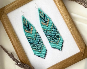 May include: A pair of long, beaded earrings with a geometric pattern in shades of turquoise, black, and gold. The earrings are hanging from a wooden frame.