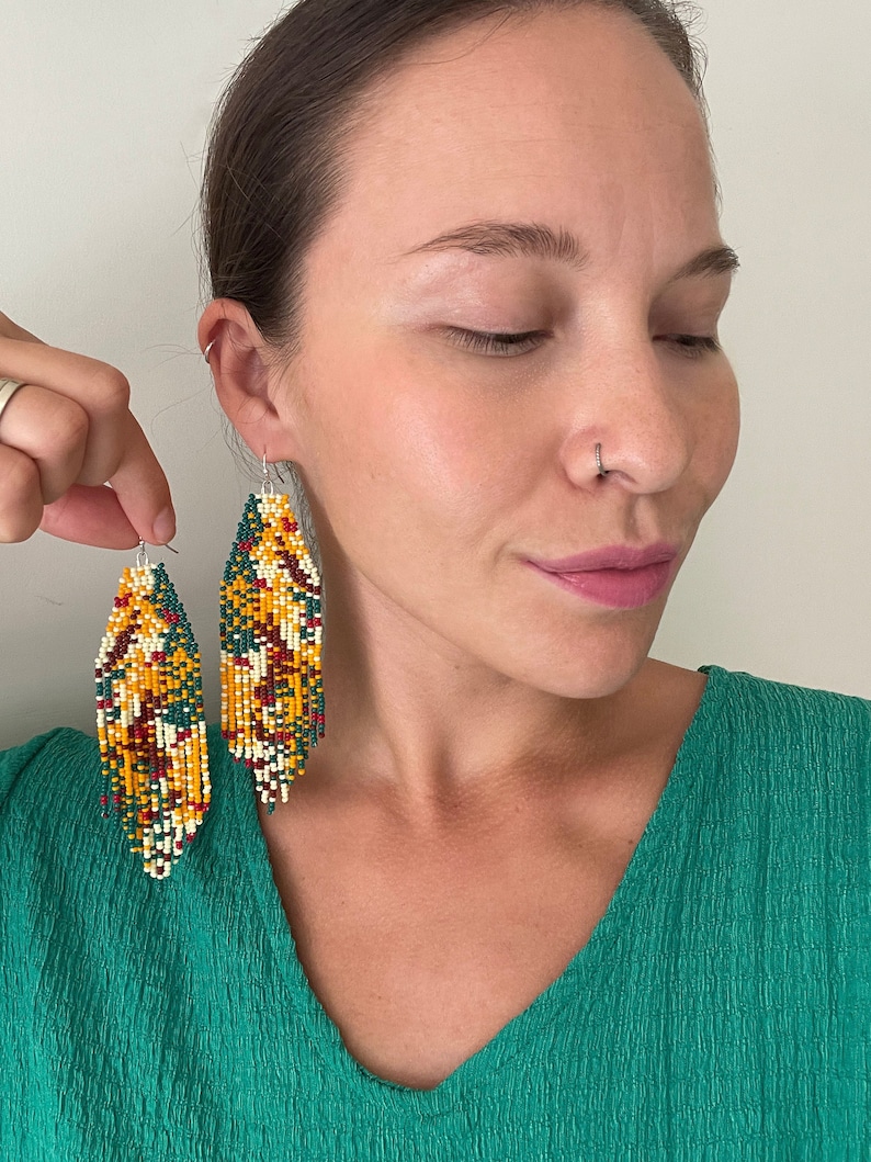 May include: A pair of long, beaded earrings with a colourful geometric pattern in shades of yellow, green, red, and white. The earrings are hanging from the ears of a person wearing a green shirt.