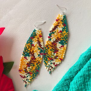 May include: A pair of long, beaded earrings with a colourful geometric pattern in shades of yellow, green, red, and white. The earrings are made of small, round beads and have a fringe-like design at the bottom.