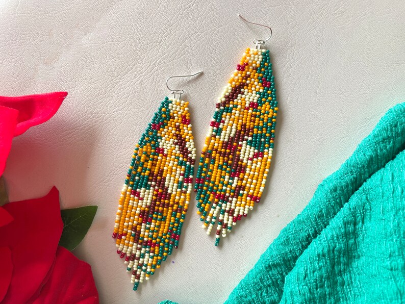 May include: A pair of long, beaded earrings with a colourful geometric pattern in shades of yellow, green, red, and white. The earrings are made of small, round beads and have a fringe-like design at the bottom.