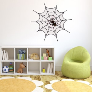 Puede incluir: Una estantería blanca con una variedad de artículos, incluyendo libros, un teléfono de juguete, un animal de peluche, una jirafa de juguete y un juego de bloques de construcción. Hay un sillón de puf verde en primer plano y una cama blanca con un edredón estampado en verde y blanco en el fondo. Una calcomanía de pared de telaraña negra con una araña está en la pared detrás de la estantería.