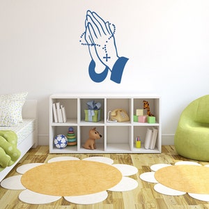 May include: A blue wall decal of praying hands with a rosary. The decal is on a white wall in a child's room with a green armchair, a white bookshelf, and a bed with a green and white comforter.