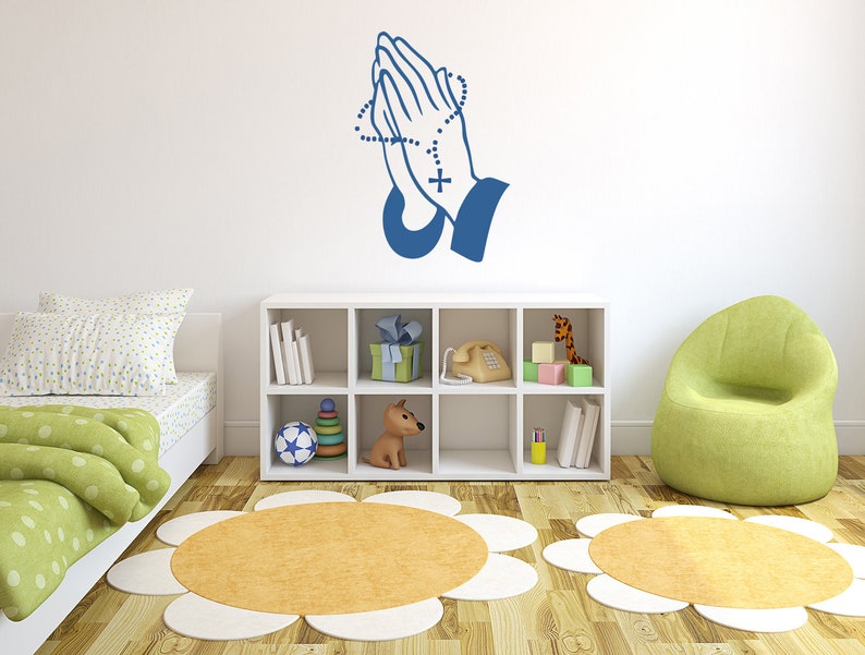 May include: A blue wall decal of praying hands with a rosary. The decal is on a white wall in a child's room with a green armchair, a white bookshelf, and a bed with a green and white comforter.