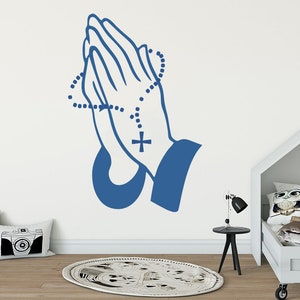 May include: Blue silhouette of two hands clasped in prayer with a cross and a beaded rosary around the wrists. The hands are on a white wall.