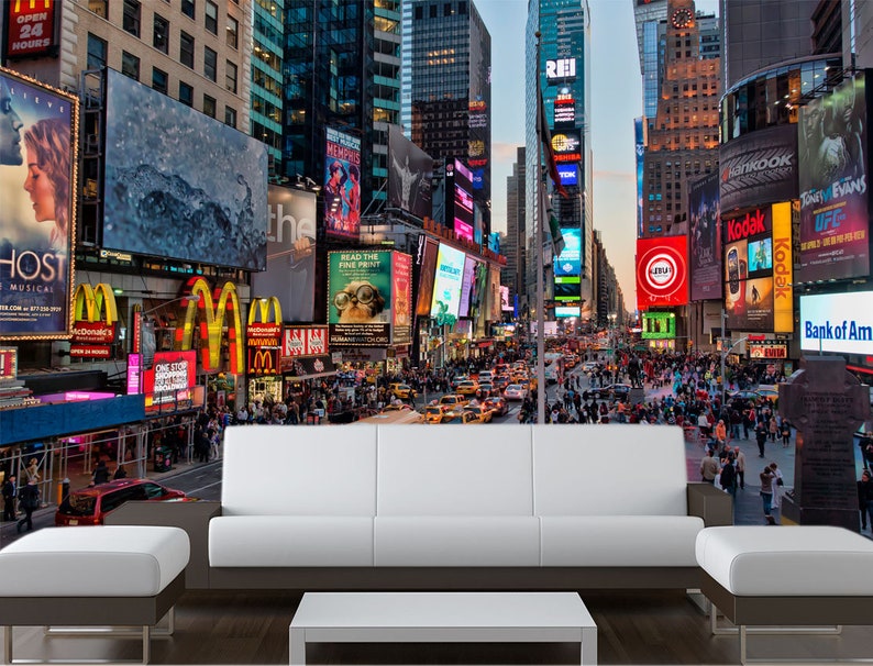 May include: A colorful cityscape mural featuring Times Square in New York City with bright lights, billboards, and a crowd of people walking on the street. The mural is displayed on a wall behind a white couch and a coffee table.