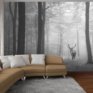 May include: A black and white photograph of a deer standing in a foggy forest. The deer is facing away from the camera and has large antlers. The forest is dense with tall trees and the ground is covered in leaves.