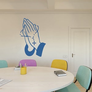 May include: A blue praying hands wall decal with a rosary and cross. The decal is on a white wall in a room with a table and chairs.
