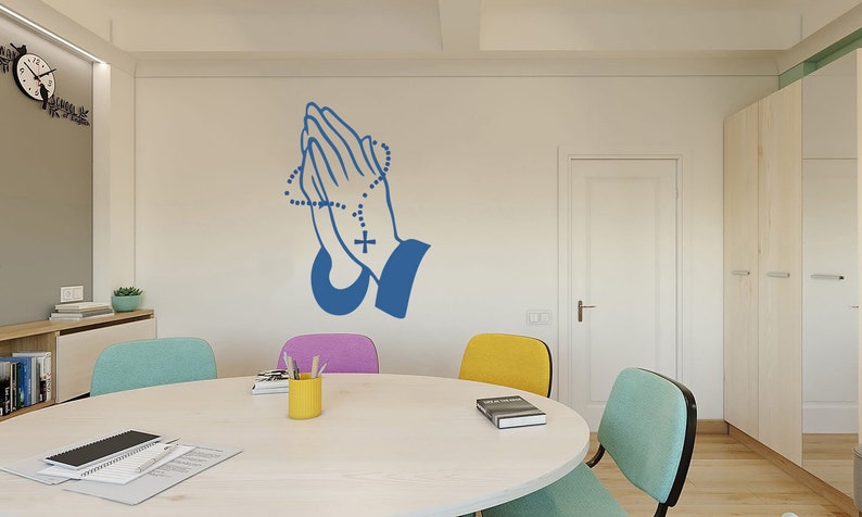 May include: A blue praying hands wall decal with a rosary and cross. The decal is on a white wall in a room with a table and chairs.