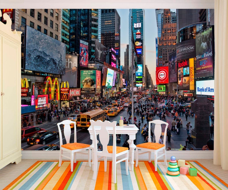 May include: A colorful photo mural depicting Times Square in New York City at dusk. The image features bright lights, billboards, and a bustling crowd of people walking on the street. There are yellow taxis and a yellow school bus in the street.