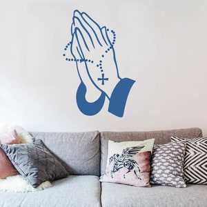 May include: A blue silhouette of two hands clasped in prayer with a rosary around the wrists. The hands are on a white wall above a grey sofa with cushions.
