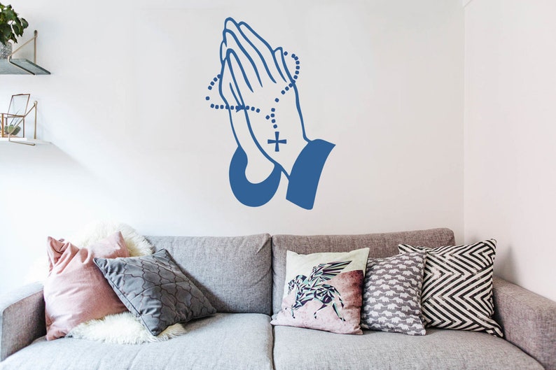 May include: A blue silhouette of two hands clasped in prayer with a rosary around the wrists. The hands are on a white wall above a grey sofa with cushions.