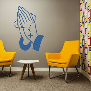 May include: Two yellow chairs with chrome legs and a small white table with wooden legs in a room with a grey wall and a colourful geometric patterned wall. A blue praying hands decal with a rosary is on the grey wall.