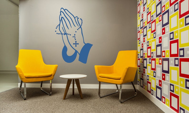 May include: Two yellow chairs with chrome legs and a small white table with wooden legs in a room with a grey wall and a colourful geometric patterned wall. A blue praying hands decal with a rosary is on the grey wall.