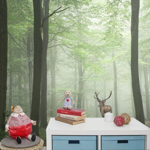 May include: A white table with two drawers sits in front of a foggy forest scene wallpaper. On the table are a stack of books, a teddy bear, a deer figurine, and two decorative balls.
