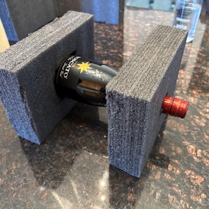 May include: A wine bottle is packaged in a gray foam box with a cut-out for the bottle. The bottle is red with a gold label that says "OLIVATO" and has a gold star on it.